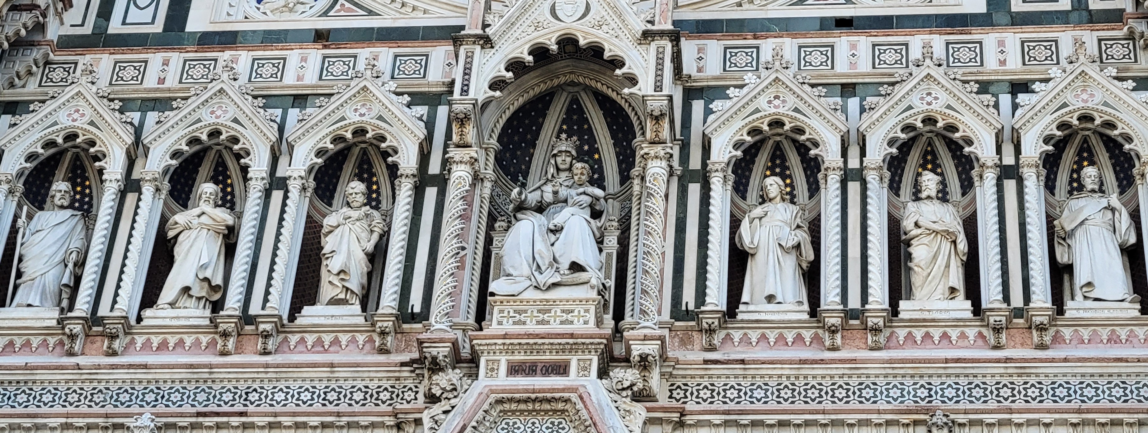 A photograph of an elaborate panel on a Catholic church that is meant to show 6 apostles and Mary in the middle, wearing
 a crown. Ignoring the time clash, on her knee is Jesus, still as a baby.